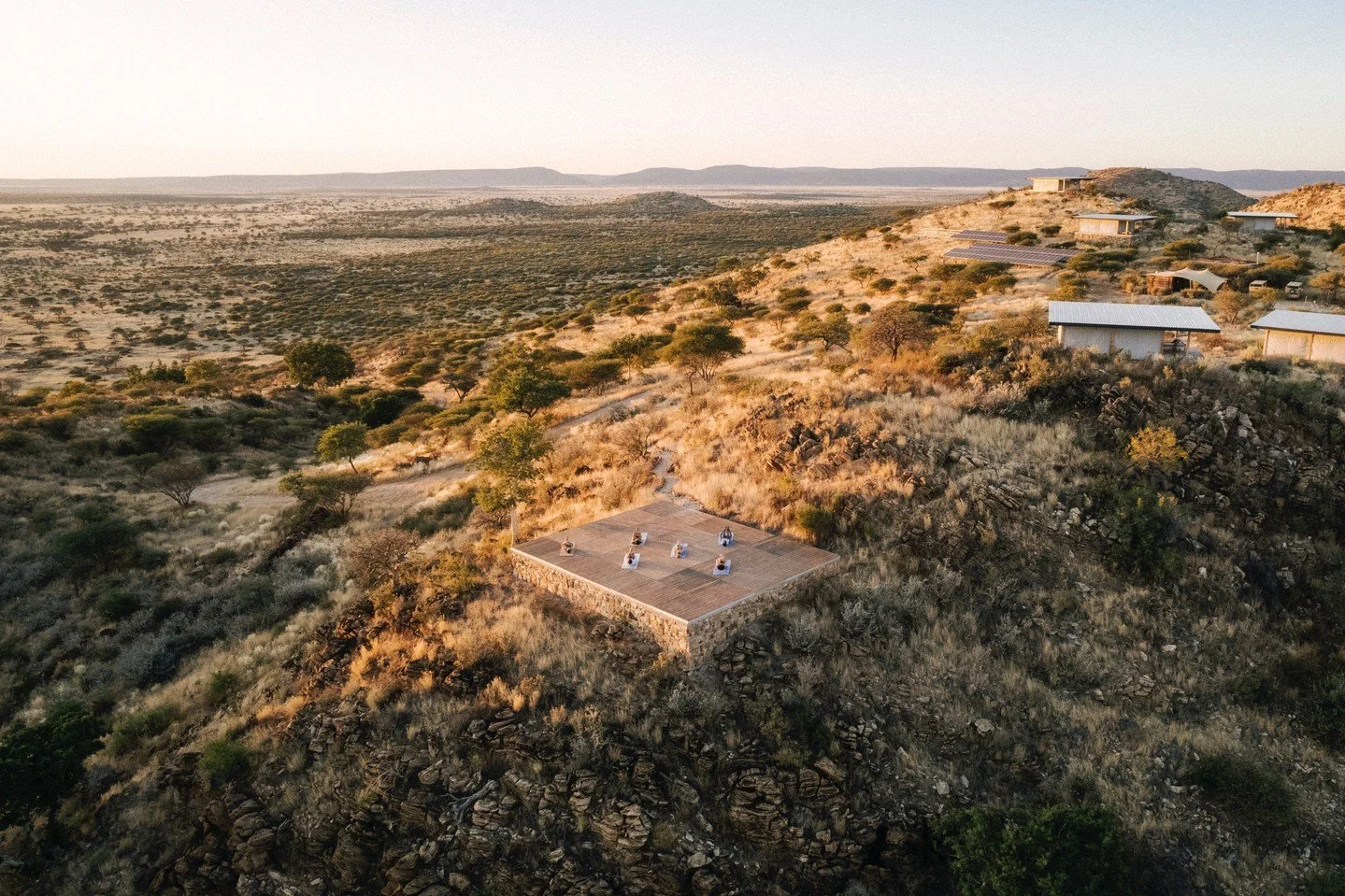 Aerial view of a desert landscape with a flat wooden deck and six sun loungers, surrounded by sparse trees and rocky terrain, with modern buildings on the hillside in the background.