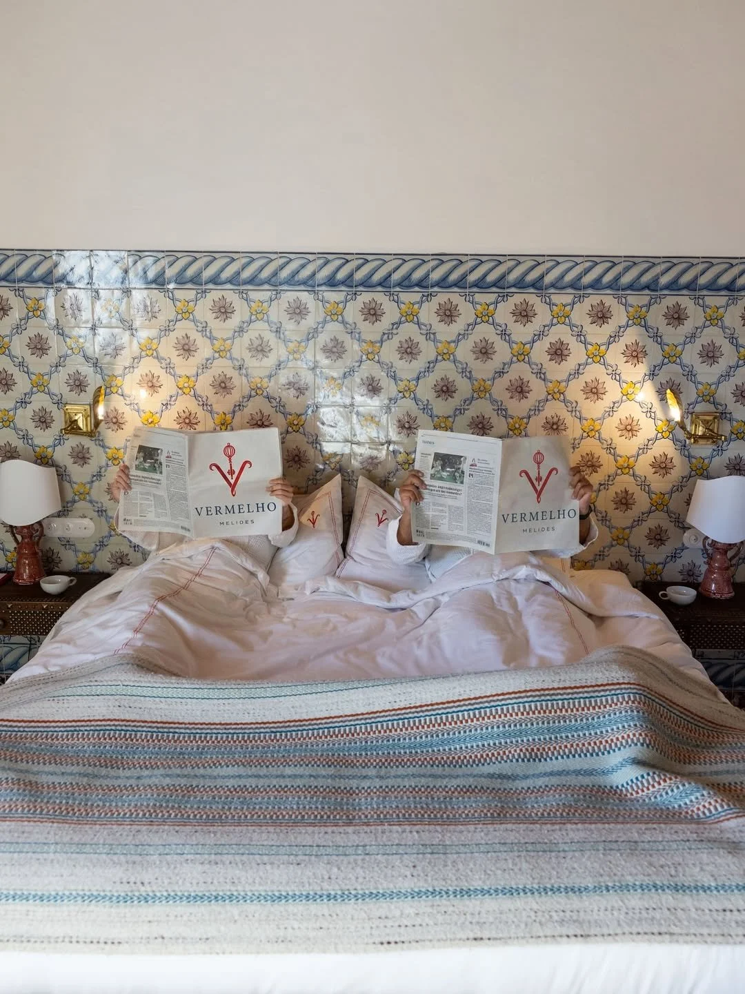 Two people lying in bed, reading newspapers titled 'Vermelho Melides', with ornate tile wall and lamps in the background.