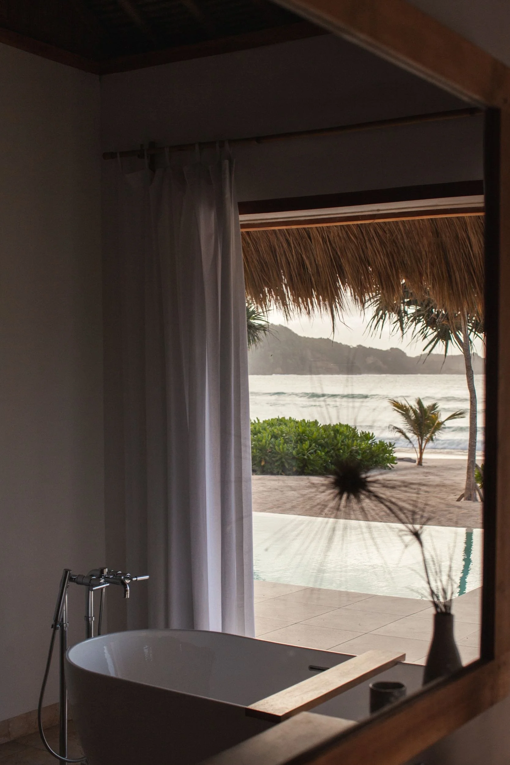 Bathroom with a white bathtub, a window with a white curtain, and a view of an outdoor tropical landscape with a thatched roof, trees, water, and mountains.