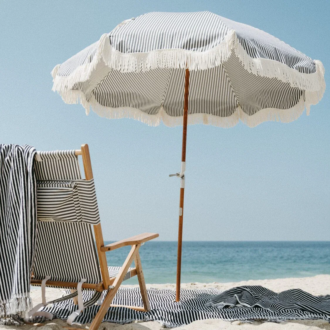 Beach scene with striped umbrella, wooden beach chair, and striped towel on sand, overlooking water
