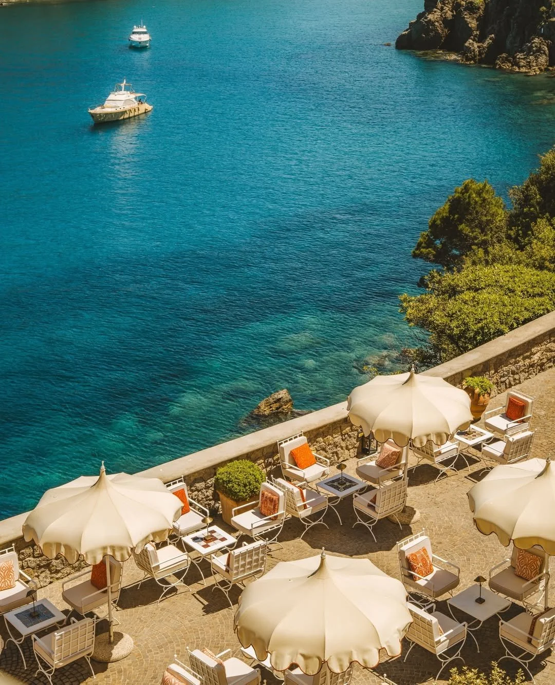 Outdoor patio with white chairs, tables, and large white umbrellas overlooking a body of water with boats and rocky shoreline.