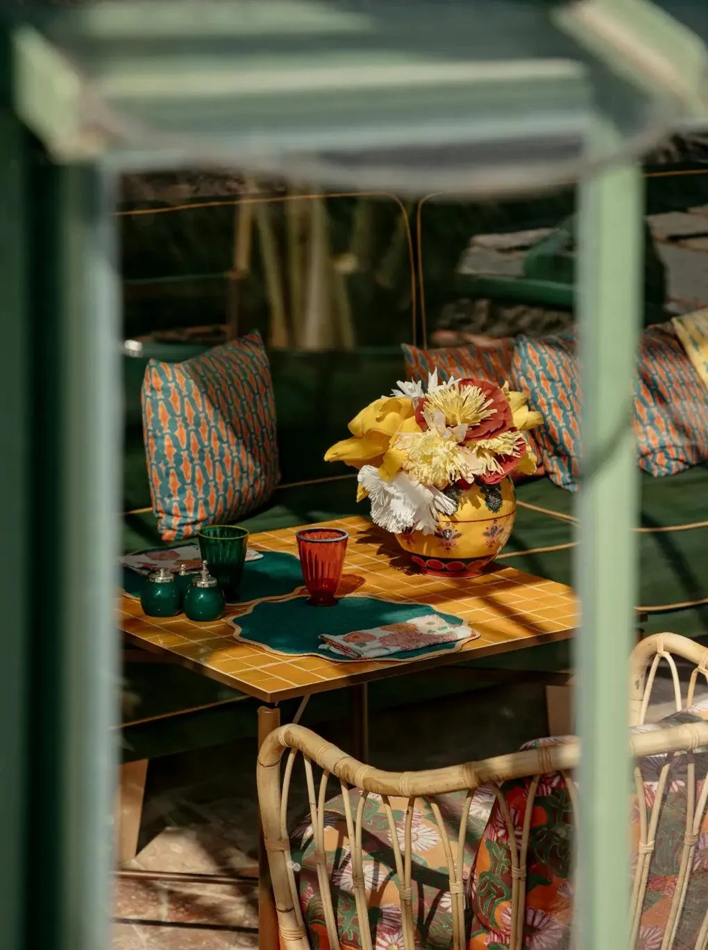 Colorful outdoor dining table with a yellow vase holding a large bouquet of yellow, white, and red flowers, surrounded by patterned cushions and colorful glassware.