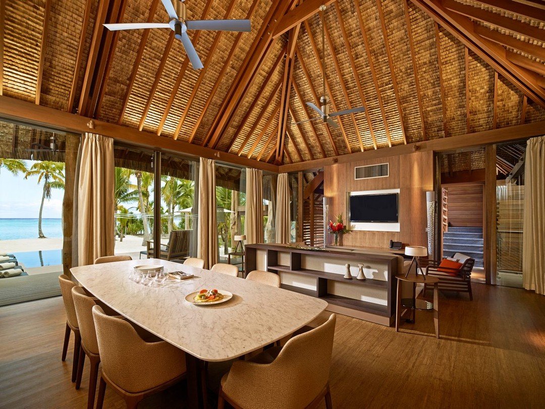 Interior view of a tropical beachfront villa with a high vaulted wooden ceiling, large sliding glass doors with curtains, overlooking palm trees and the ocean. Dining table with eight chairs, a TV on the wall, and beachside seating visible outside.