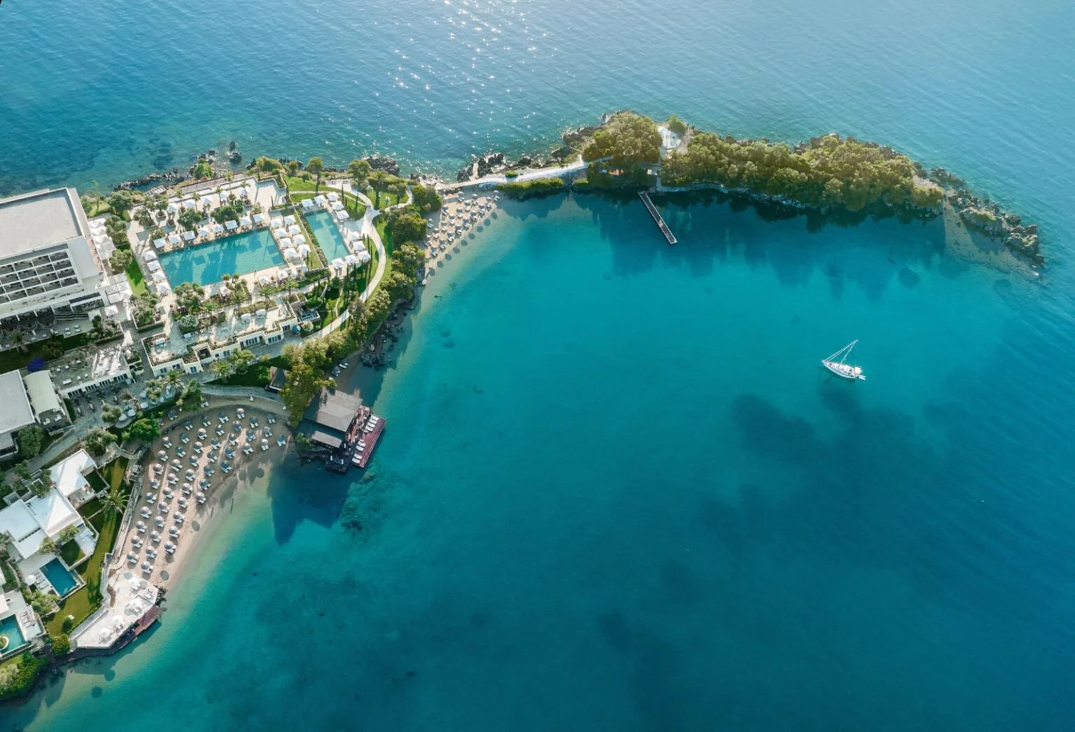 Aerial view of a resort with a large swimming pool, sun loungers, and a beach with umbrellas, next to a body of water with a sailboat, near a small island with trees.