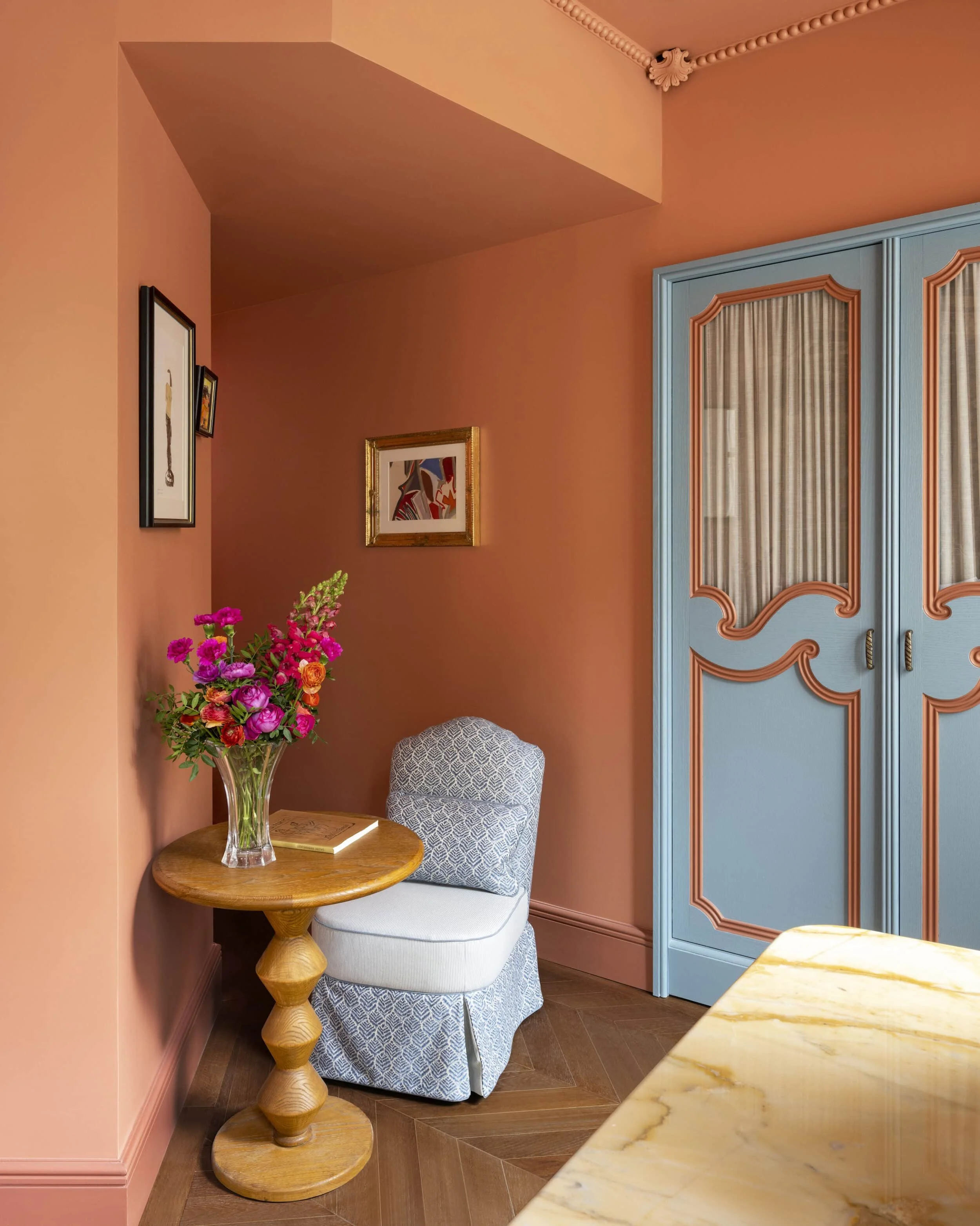 A corner of a room with pink walls and a ceiling, decorated with framed artwork. A small wooden side table holds a glass vase with pink and orange flowers. Next to the table, there's a patterned armchair with a light-colored fabric. A blue cabinet with decorative orange trim and curtains obscuring the interior is visible on the right side. A marble countertop is in the foreground.