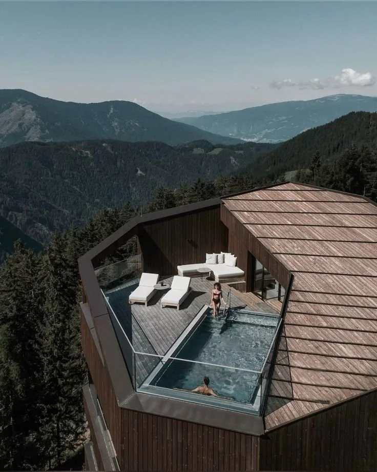 A rooftop pool with glass sides on a wooden deck, surrounded by mountain scenery and trees.