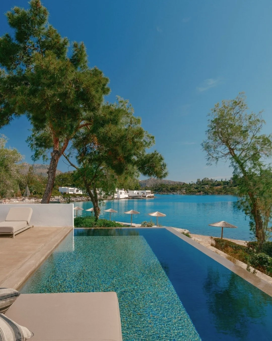 A poolside view overlooking a lake with trees, white buildings, and umbrellas under a clear blue sky.