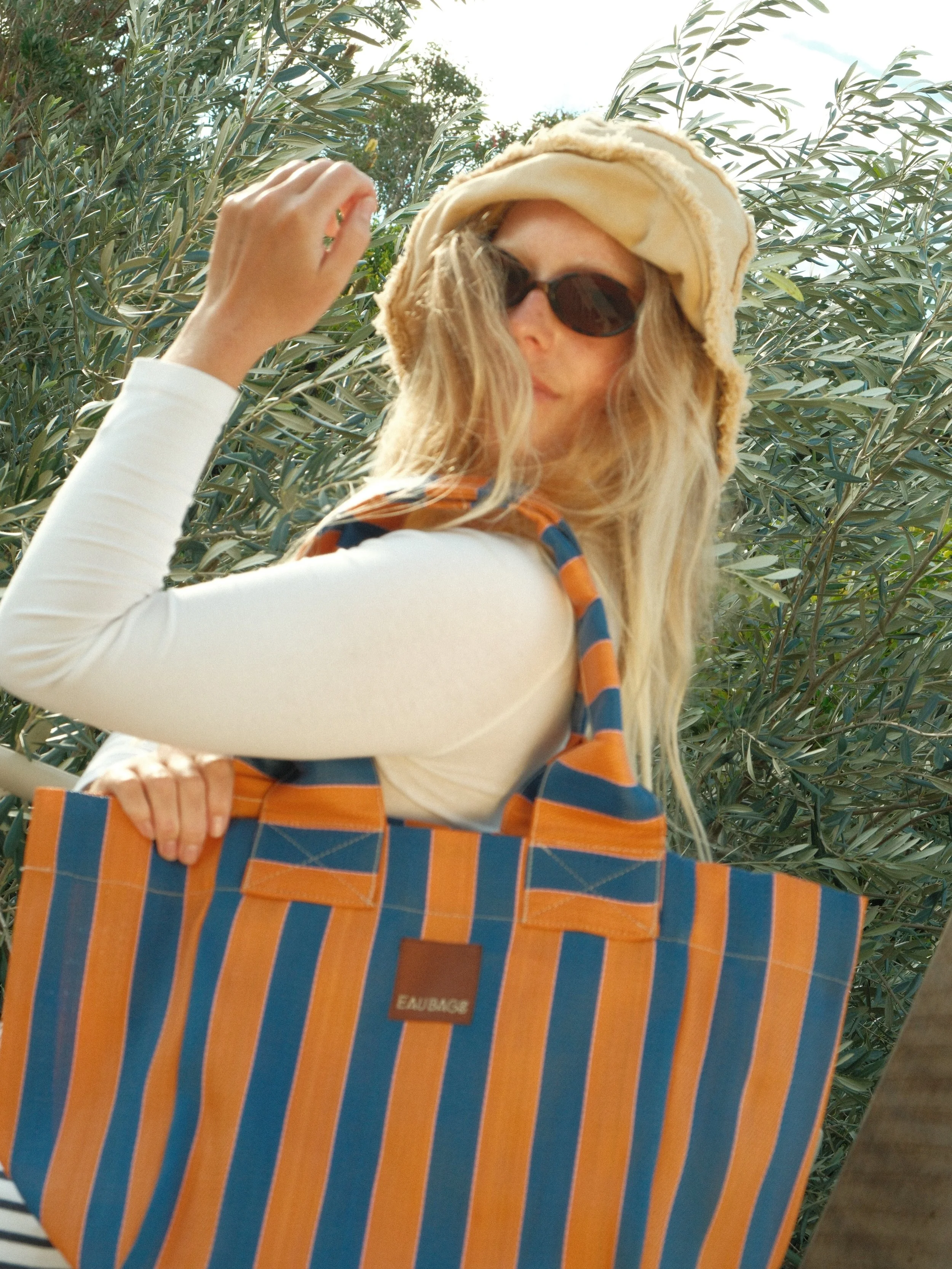 A woman wearing sunglasses, a beige hat, a white top, and a multicolored striped bag posing outdoors with green foliage in the background.