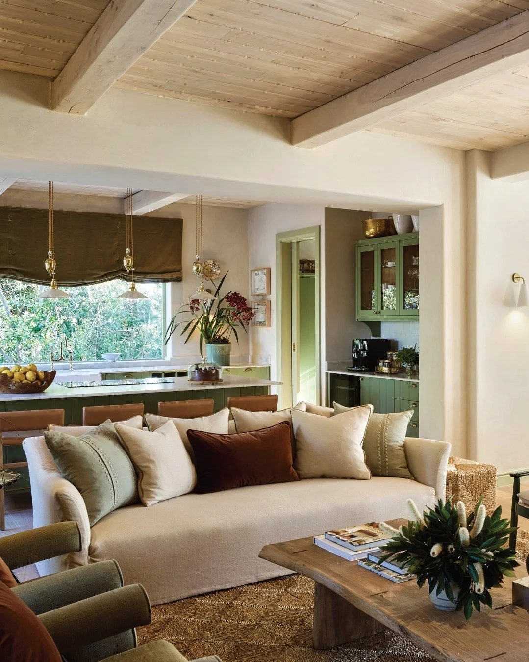 Cozy living room with beige sofa, multiple pillows, wooden coffee table with a flower arrangement, green kitchen cabinets, and large window with greenery outside.