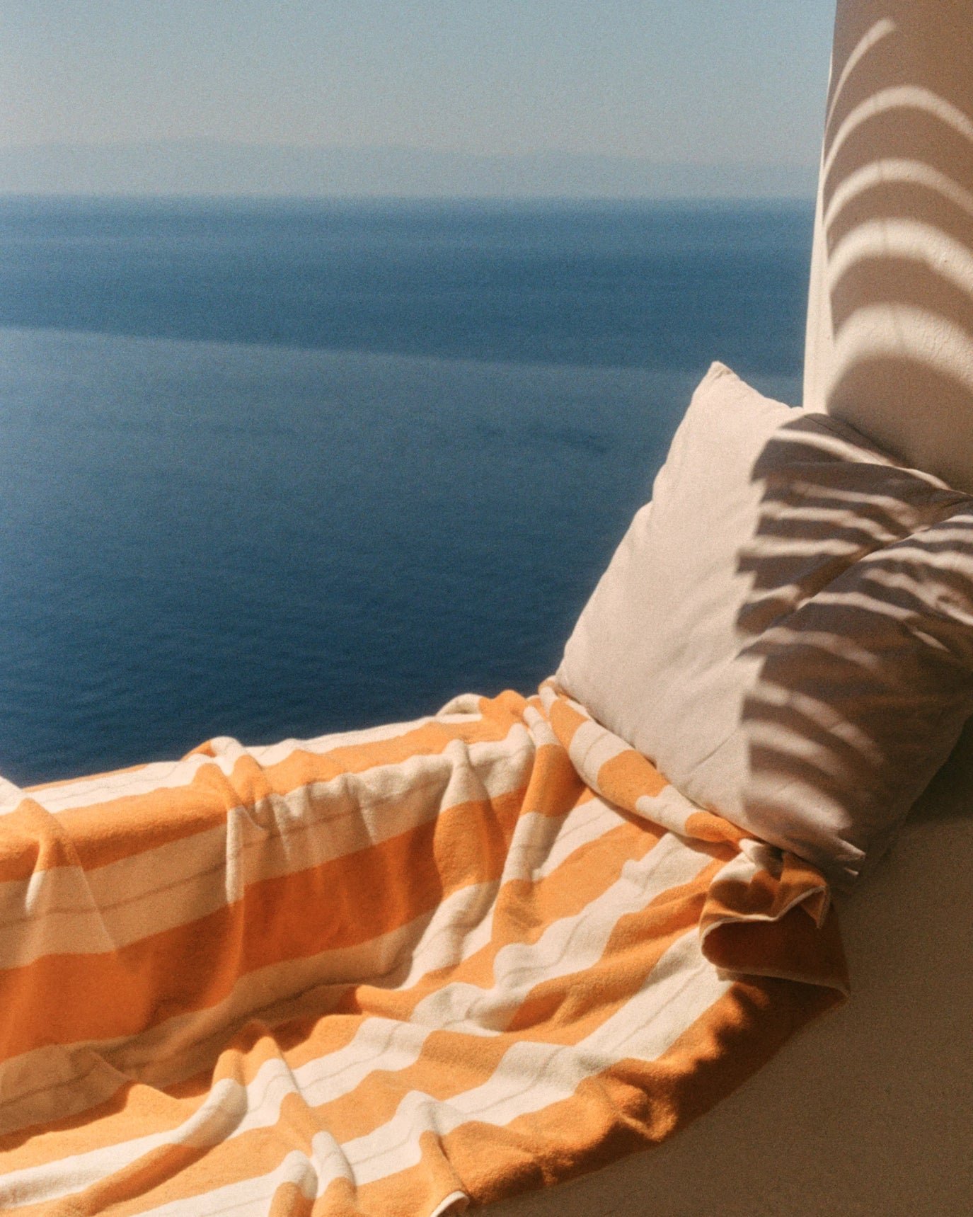 view of a bed with orange and white striped sheets, pillows, and a towel in the foreground, with a wall and ocean visible in the background