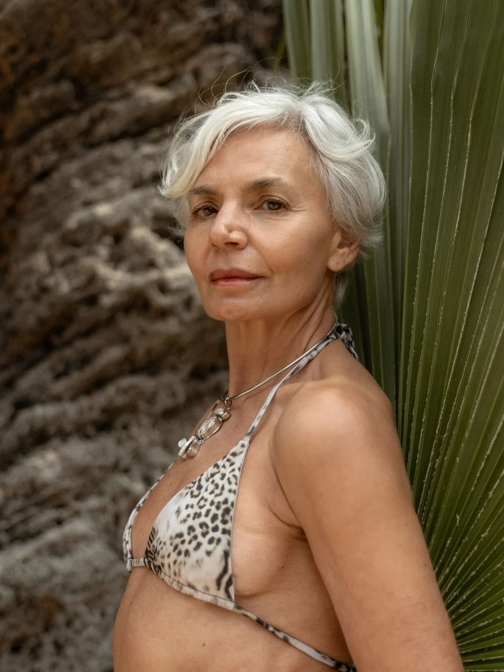 An elderly woman with short, wavy gray hair stands in front of a textured background with green palm leaves on one side, wearing a leopard print bikini top and a necklace.