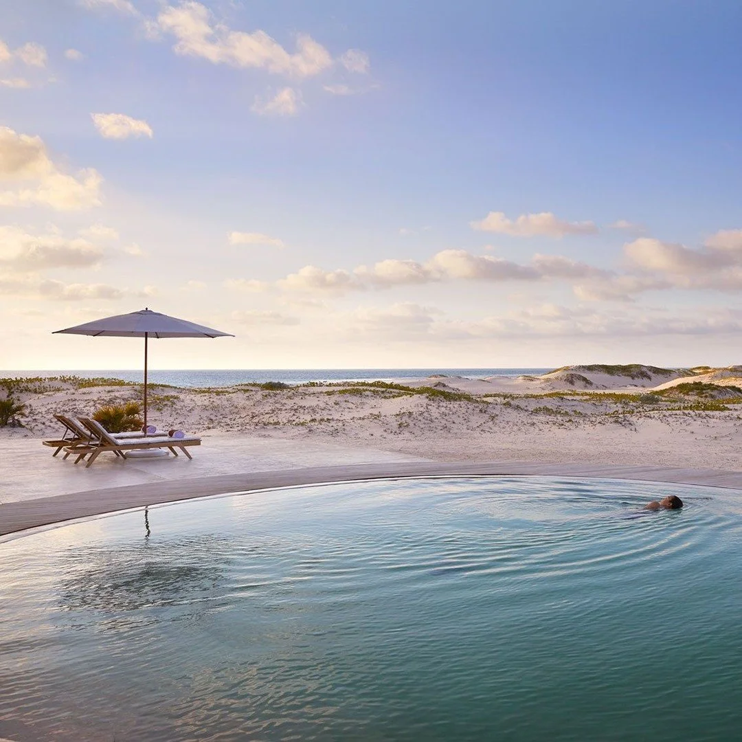 A person swimming in an outdoor pool on a sandy beach with a blue sky and clouds, lounge chairs, and an umbrella nearby.