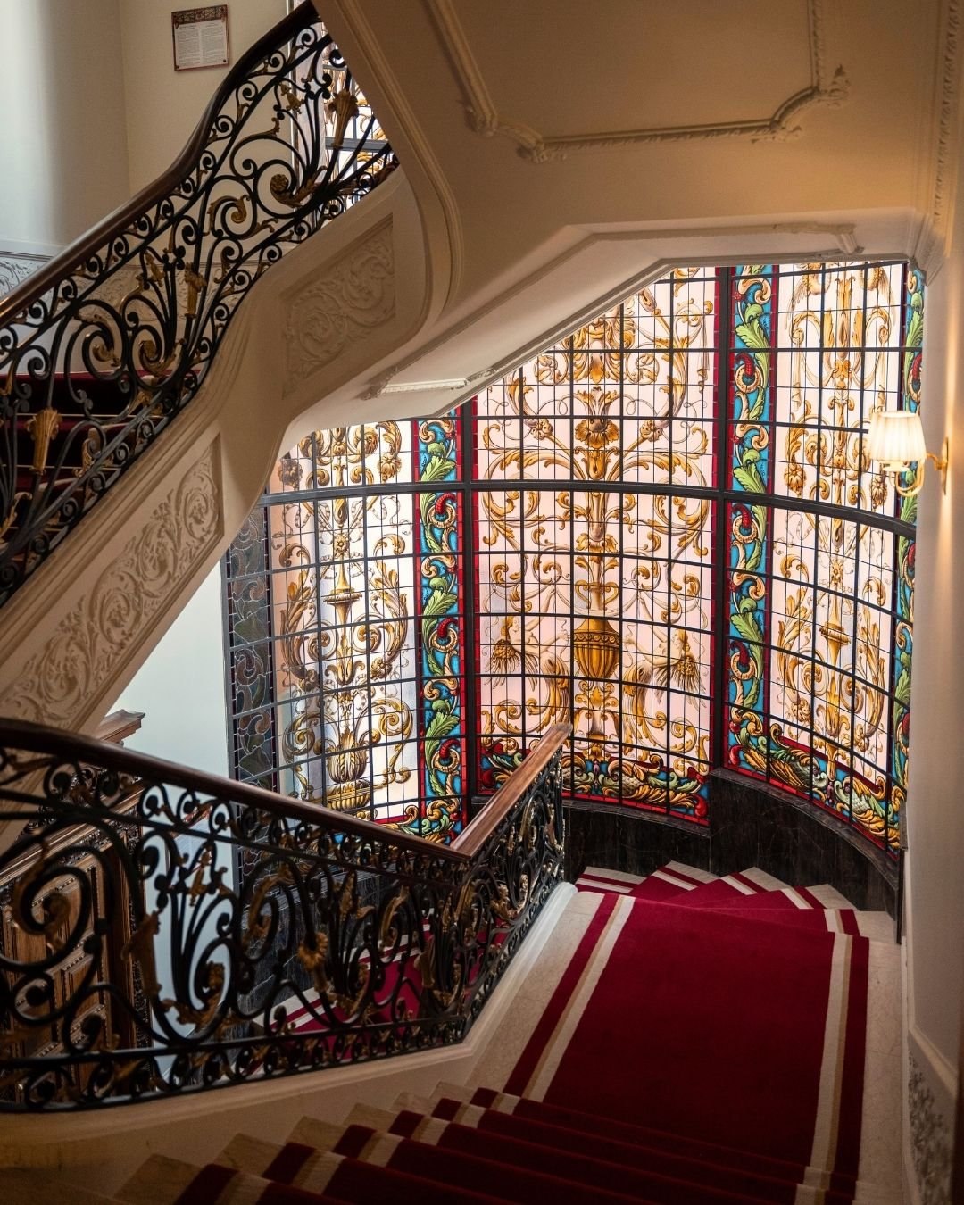 Elegant staircase with ornate black and gold wrought iron railing, red and white striped carpet, and a large stained glass window with gold, blue, green, and red decorative patterns.