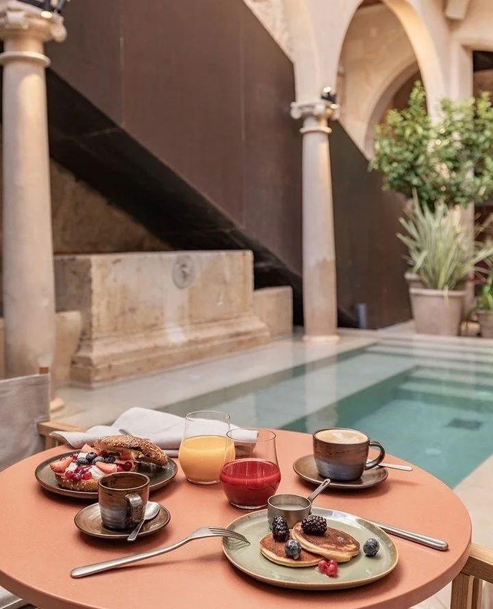 A breakfast spread on a peach-colored table near a pool indoors, including pancakes with berries, eggs, coffee, juice, and fruit.