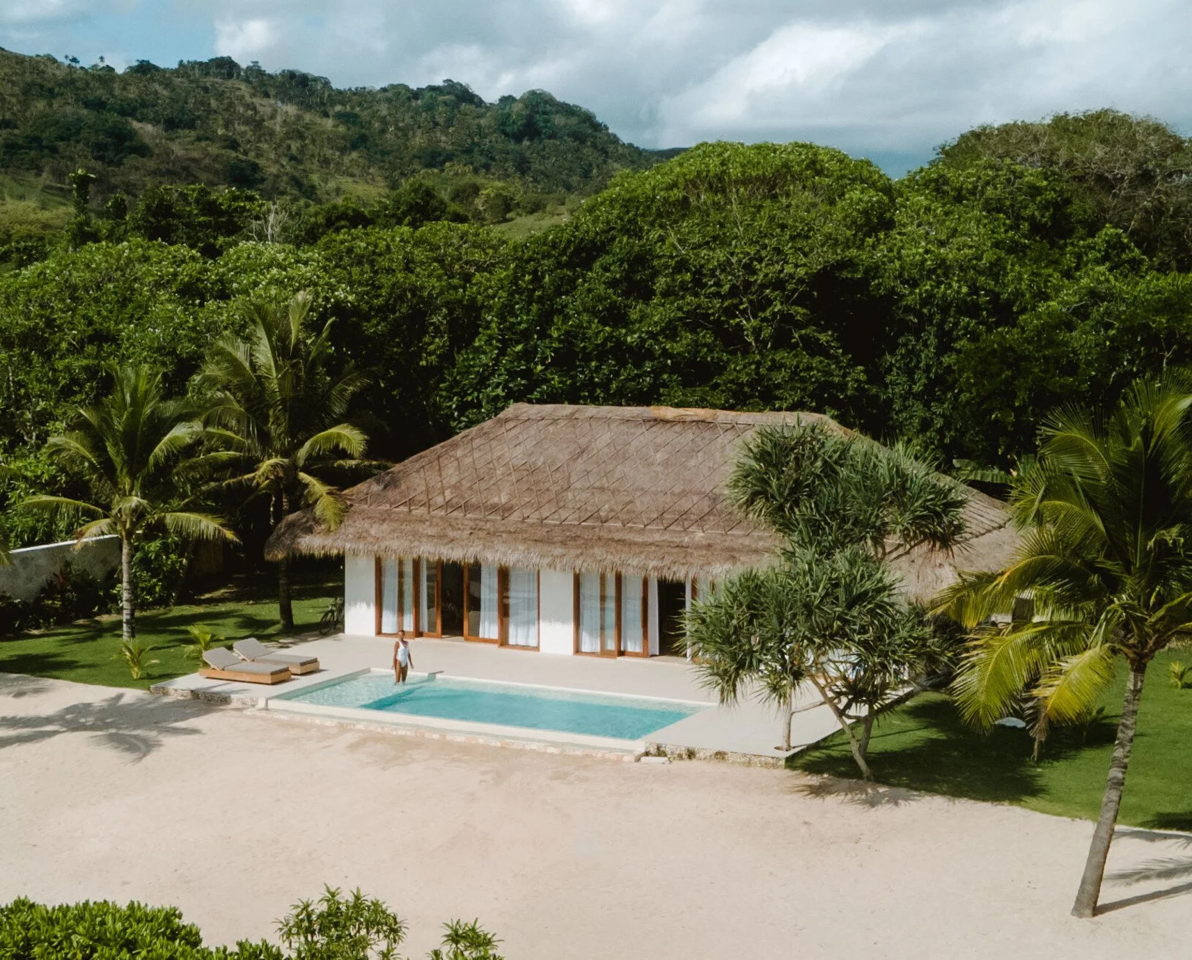 A tropical villa with a thatched roof, swimming pool, lounge chairs, and lush green trees, set against a backdrop of green hills and cloudy sky.