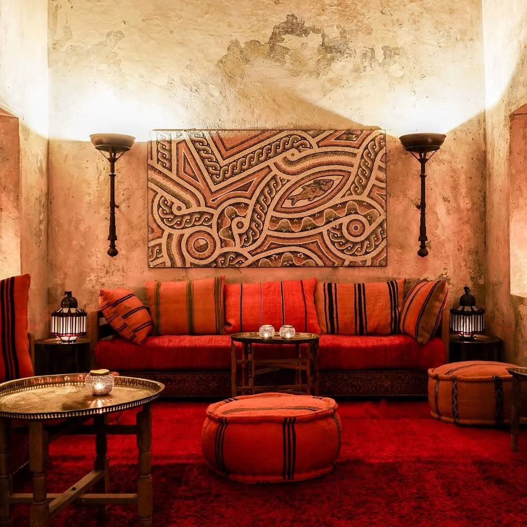 A cozy, dimly lit living room with rustic, textured walls. A large, patterned artwork hangs above a red and orange striped couch with matching pillows. Two wall-mounted sconces light the room, and small side tables with candles and lanterns are placed beside the couch. A round ottoman sits in front of the couch, and a wooden coffee table with a candle on it is in the foreground.