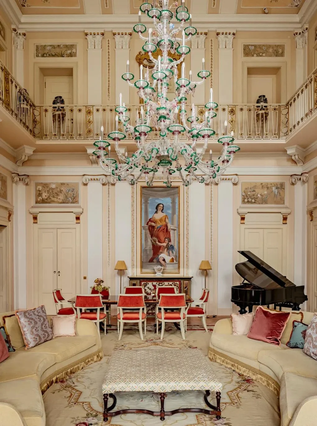Elegant living room with a grand chandelier, cream-colored walls with decorative moldings, a large portrait, a black grand piano, two beige sofas with decorative pillows, a patterned coffee table, and a dining area with red chairs and a table in the background.