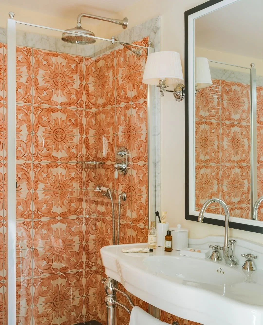 A bathroom featuring a shower with orange patterned tiles, a large mirror, a white sink with silver fixtures, and various toiletries.