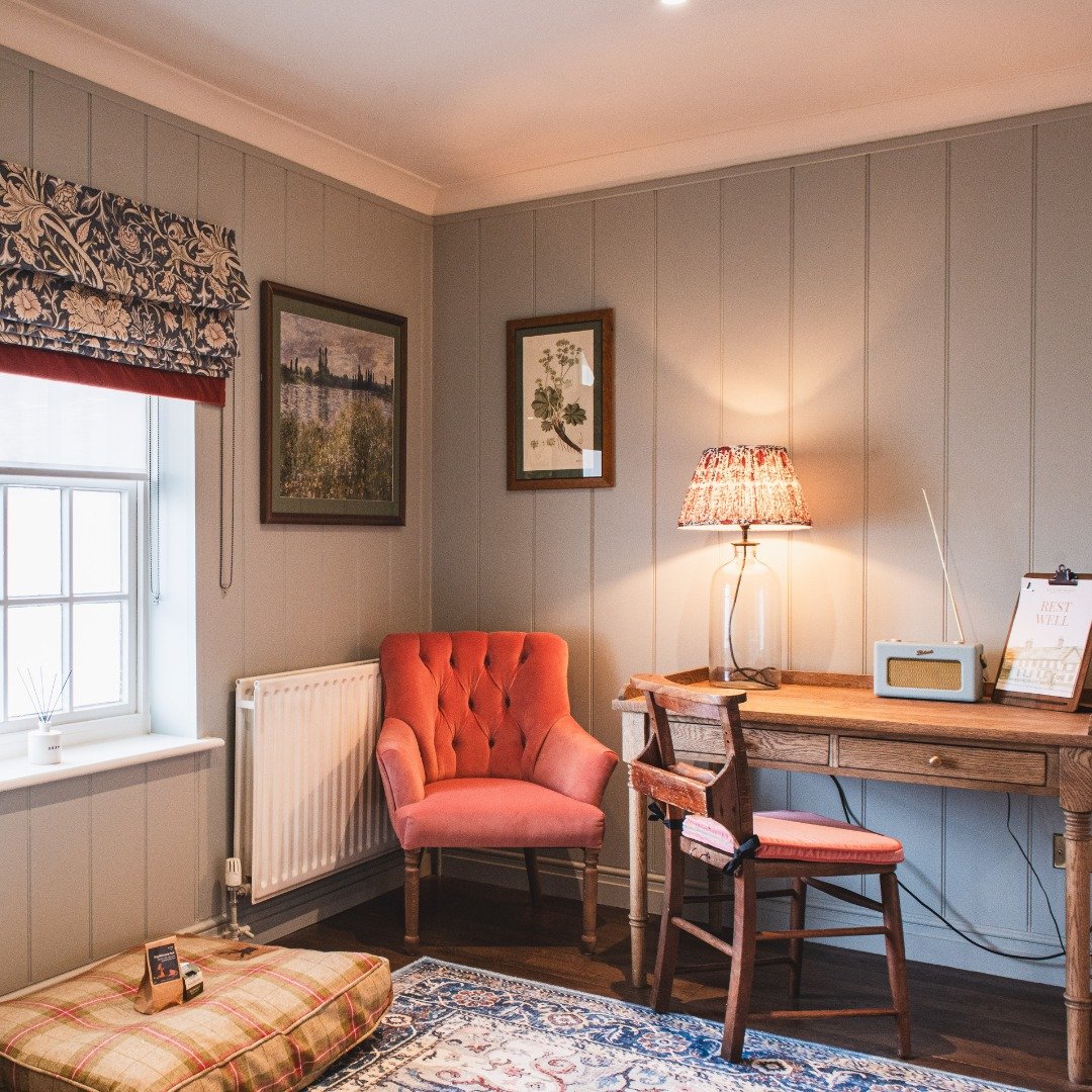 Cozy room with gray wood-paneled walls, a window with floral valance, coral armchair, wooden desk with a lamp and radio, framed pictures, and a plaid pet bed on a patterned rug.