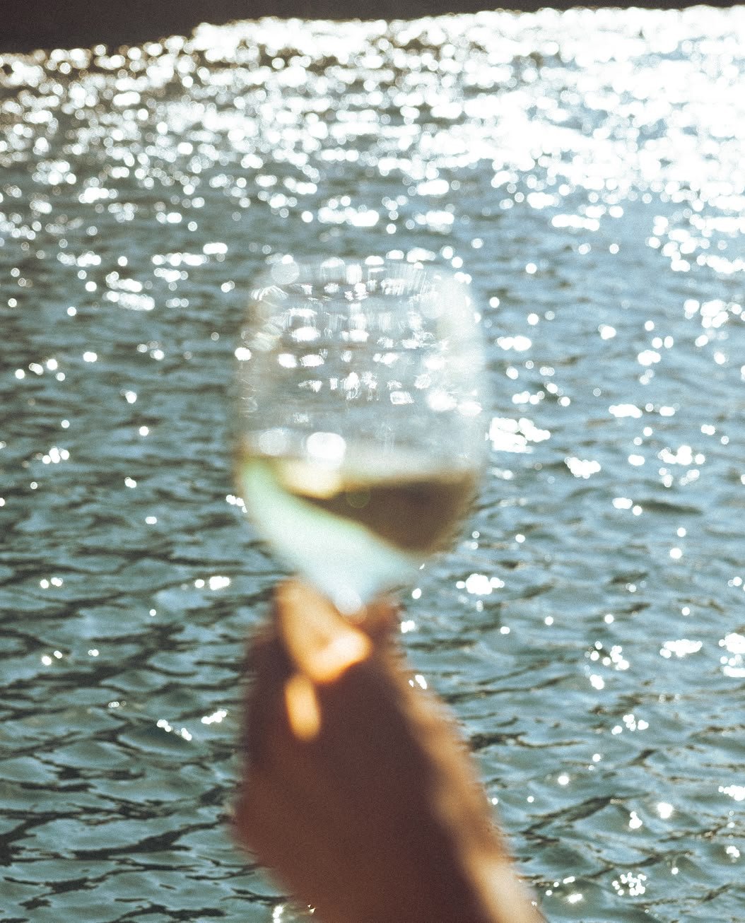 A blurry hand holding a glass of wine against a sparkling water background.