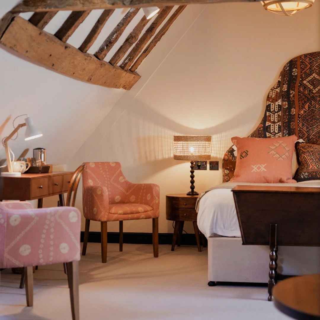 A cozy bedroom corner with a bed, pink and patterned pillows, a bedside table with a lamp, and a pink armchair. There is a wooden slanted ceiling with exposed beams and a small desk with a white lamp and coffee setup.