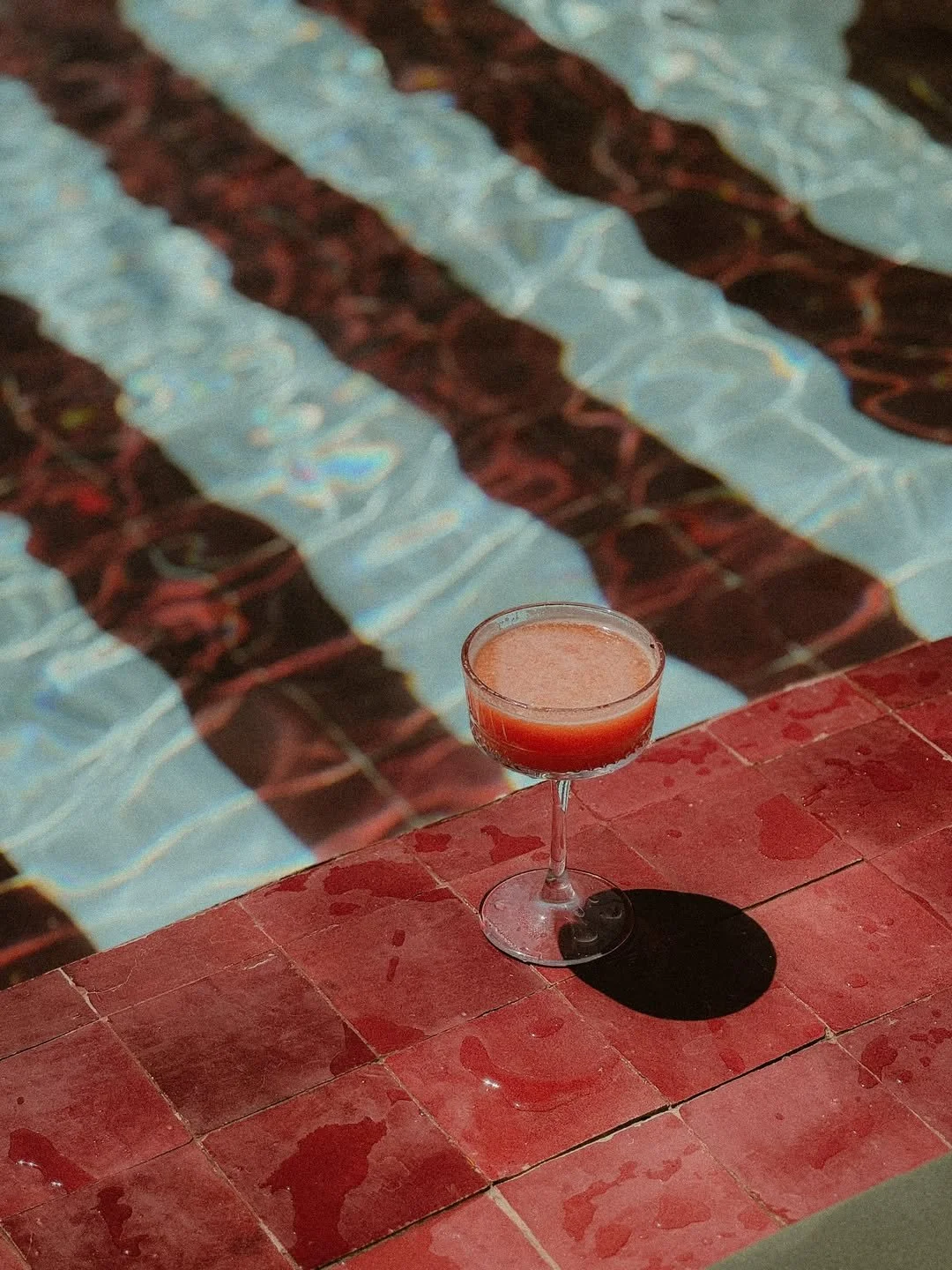 A brunch cocktail in a coupe glass on a red tiled surface next to a swimming pool with a pattern of tiles reflecting sunlight.