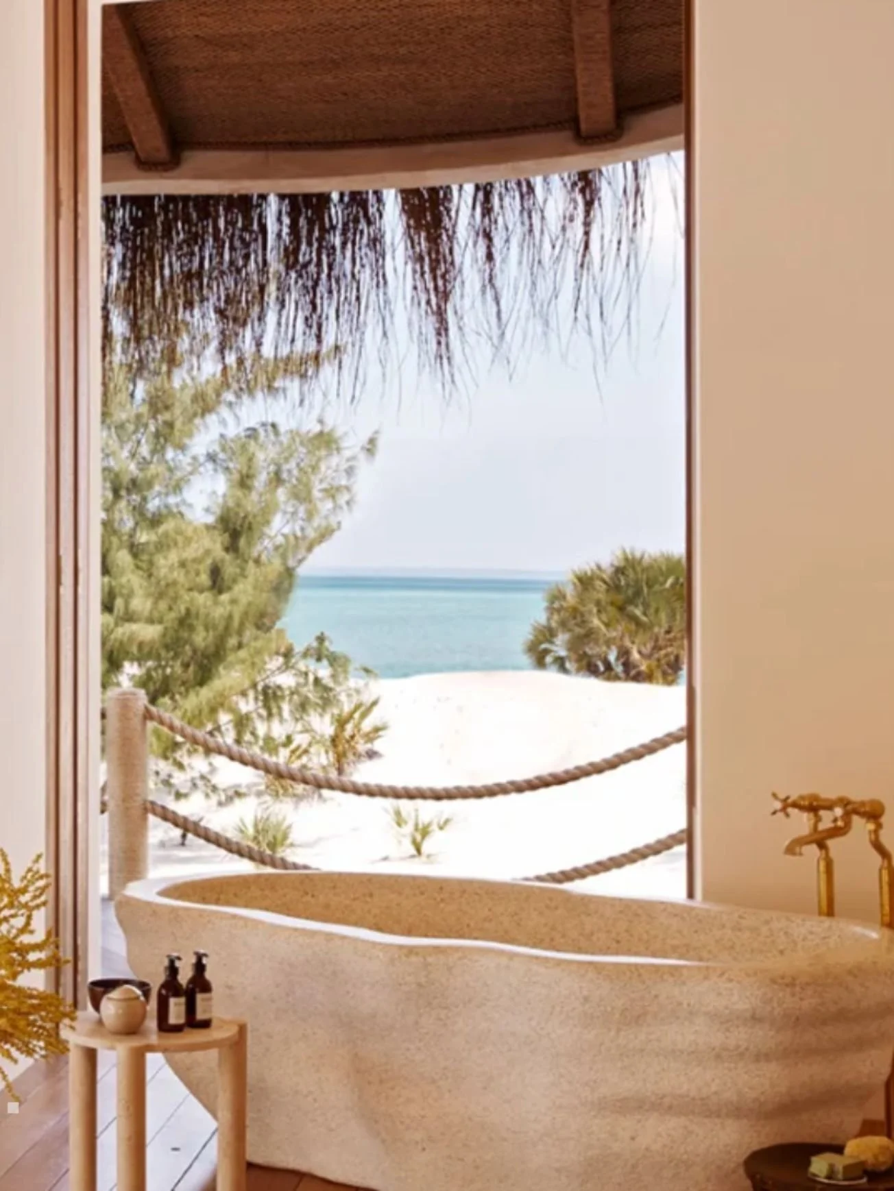 Beach view seen from a room with an opening showing sandy area, trees, and the ocean in the background. The room has a rustic bathtub, a small beige table with bottles, and rustic fixtures.