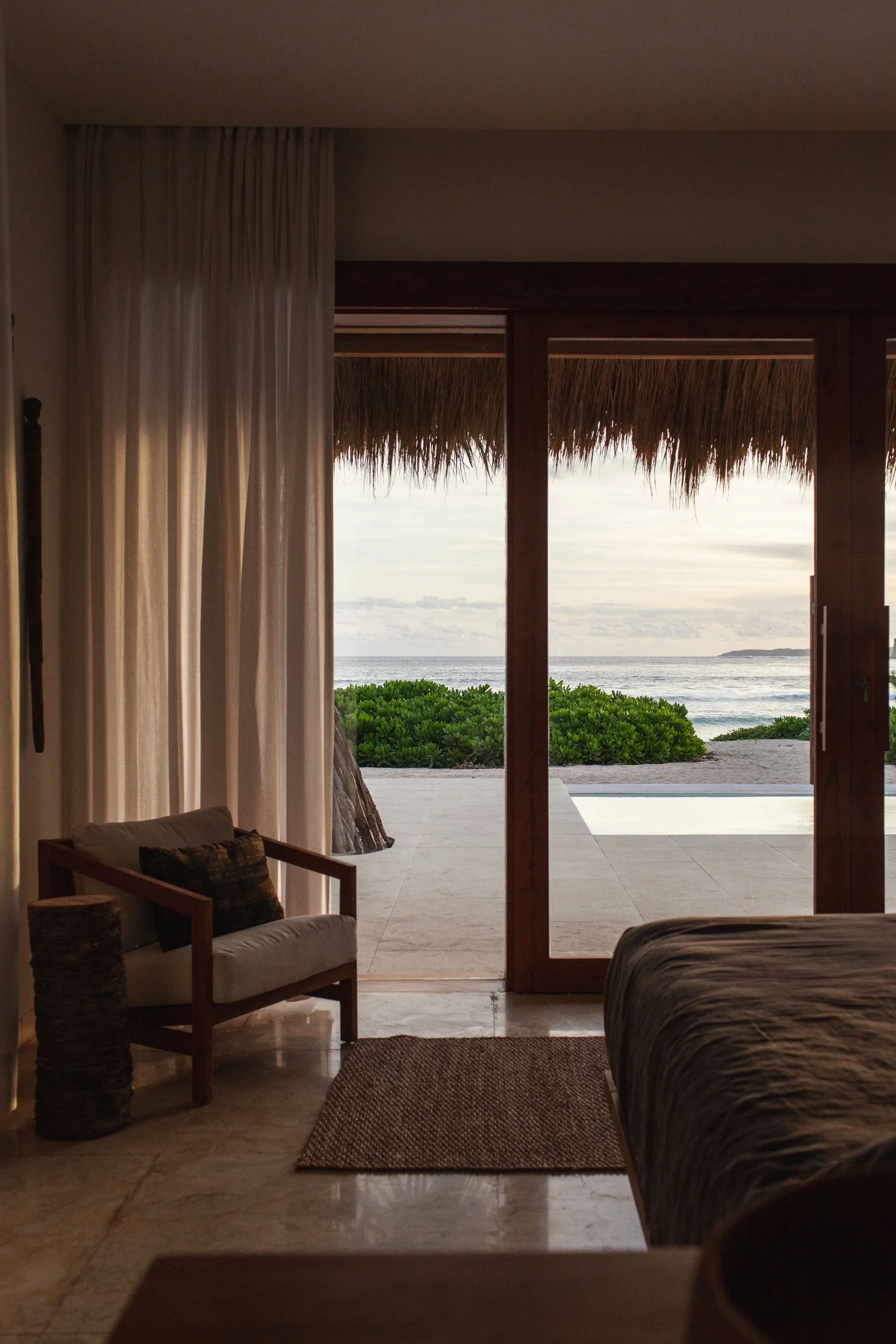 View from inside a room looking out through open sliding doors to a beach scene with sand, green bushes, and the ocean under a cloudy sky.