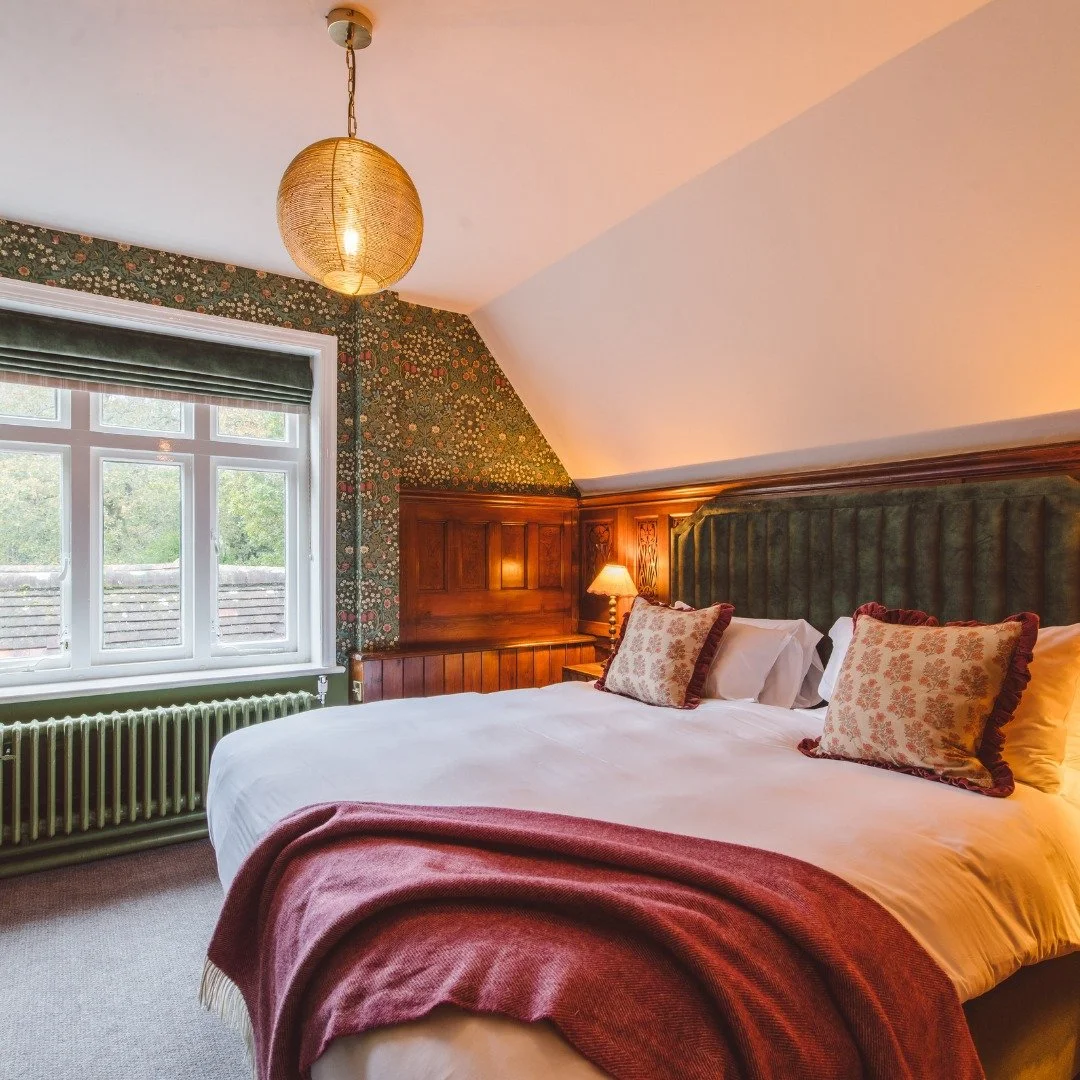 Cozy bedroom with a large bed, decorative pillows, wooden headboard, floral wallpaper, window with greenery outside, radiator, bedside lamp, and hanging ceiling light.
