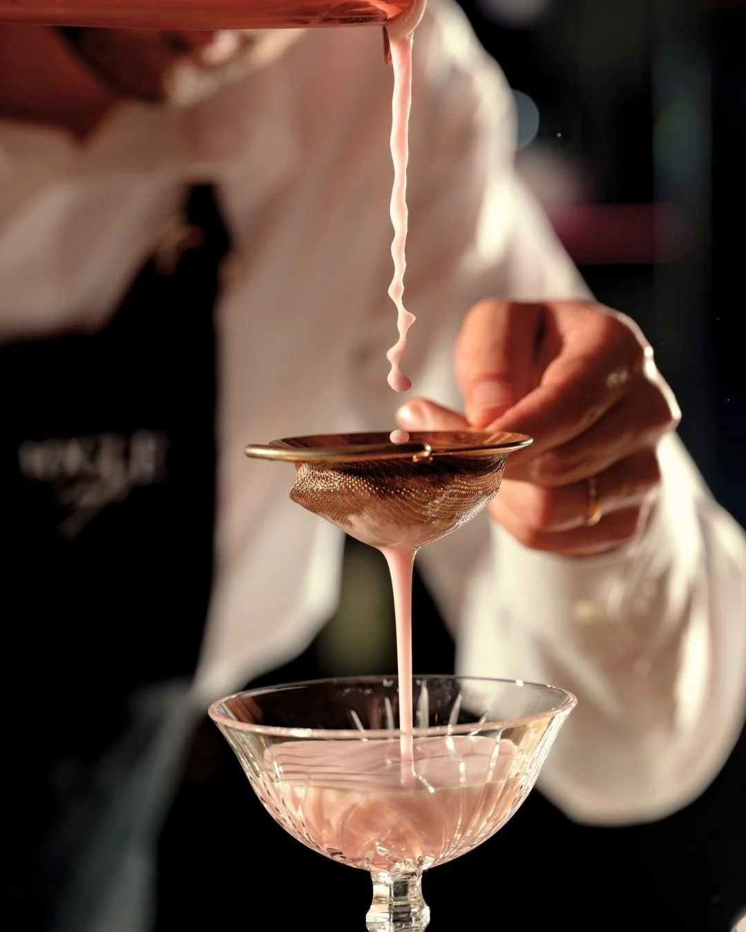 A person using a strainer to pour pink liquid into a glass coupe.