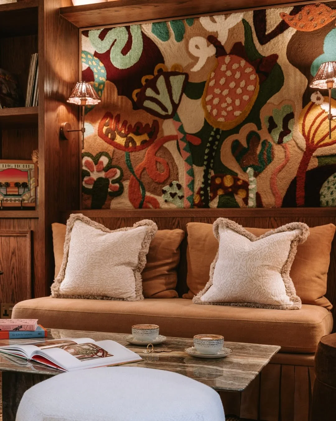 Cozy living room with a beige couch, white and tan pillows, a wooden coffee table with teacups, and a colorful abstract tapestry on the wall.