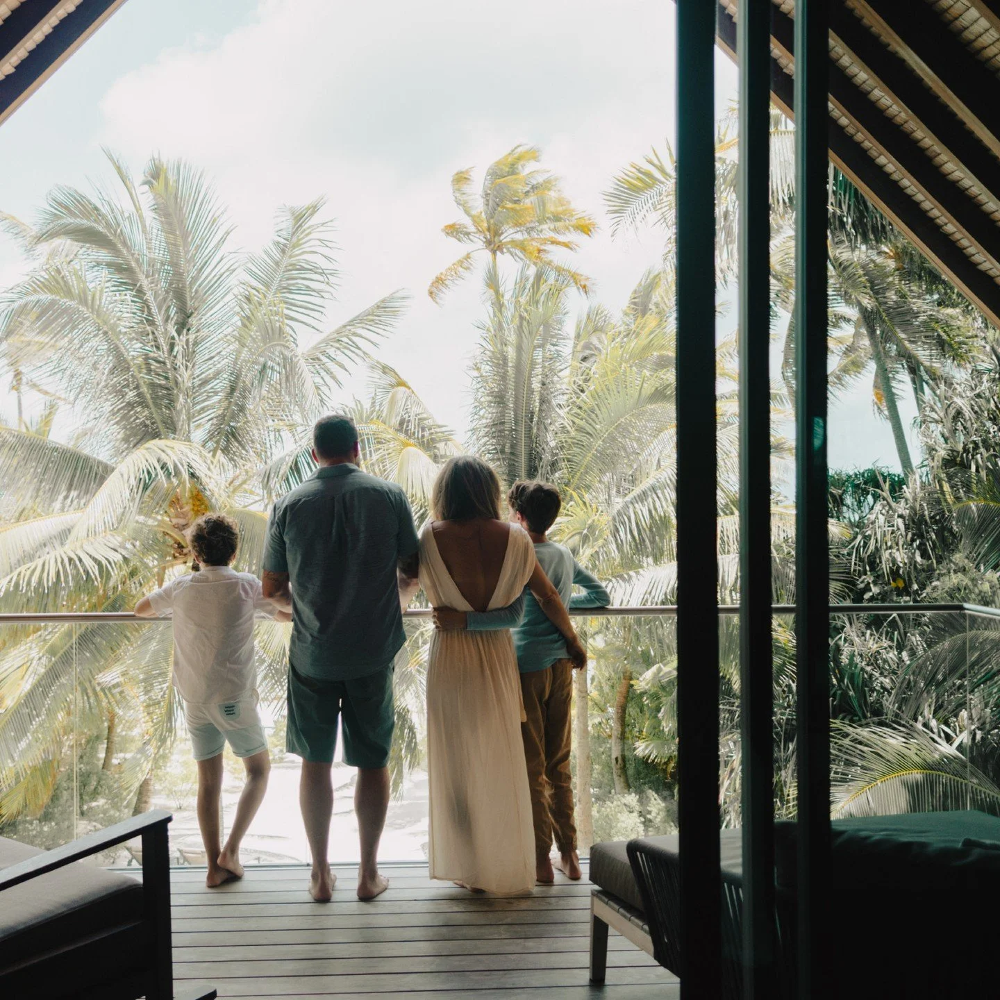 A group of five people, including two children and three adults, are standing on a balcony overlooking a tropical landscape with tall palm trees.