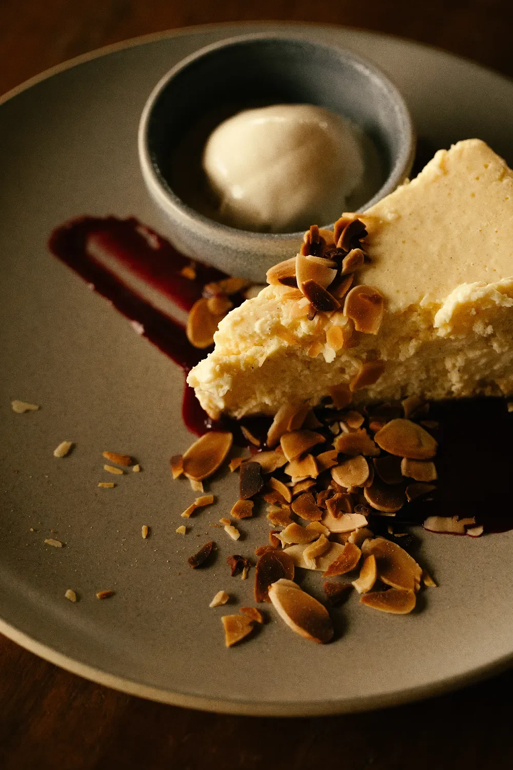 A slice of cheesecake topped with chopped almonds on a gray plate, with a dollop of whipped cream in a small black bowl and a thin streak of berry sauce on the plate.