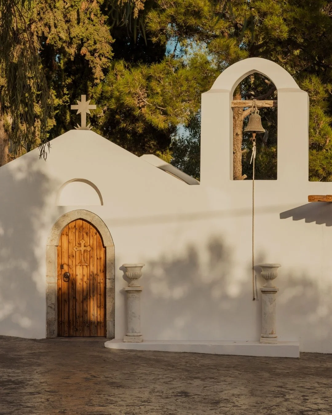 A white stucco church facade with a wooden door topped with a small cross, a bell in an arched structure, and two decorative columns, with greenery and trees in the background.