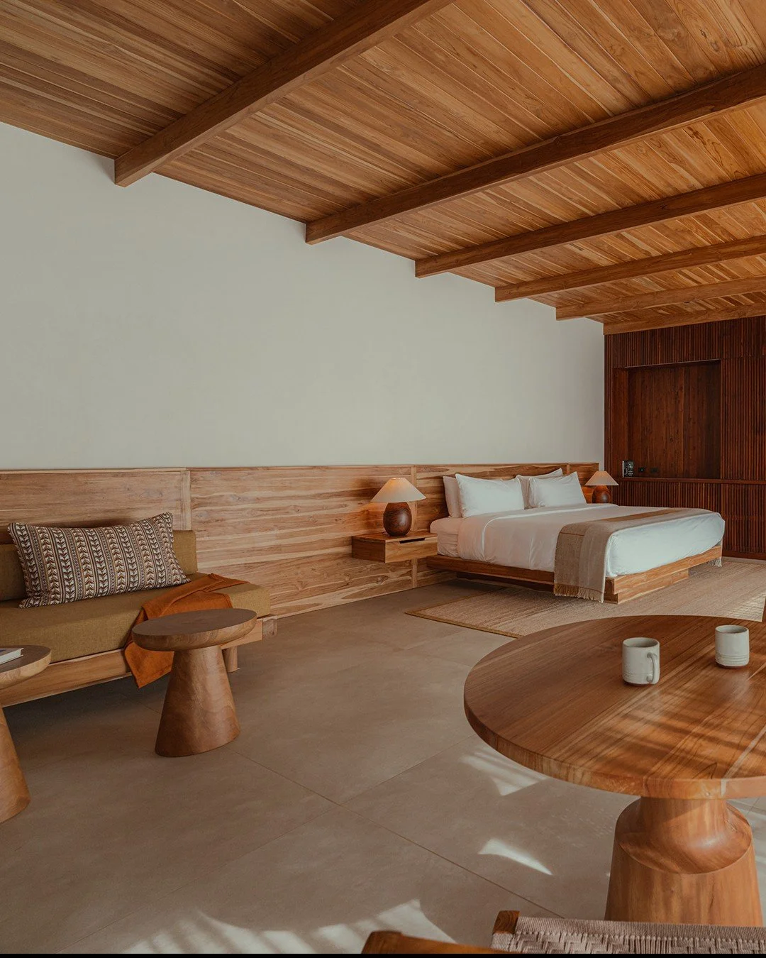 A bedroom with a wooden ceiling, a bed with white linens, two bedside lamps, a sitting area with a sofa and a round wooden table with two mugs, and beige tiled flooring.