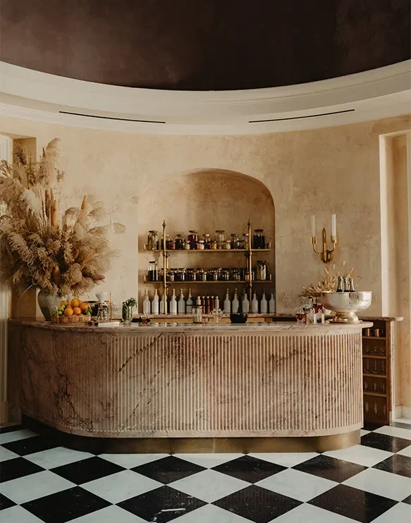 Elegant bar with a curved marble counter, shelf with bottles and jars, floral arrangement, candle sconces, and a black-and-white checkered floor