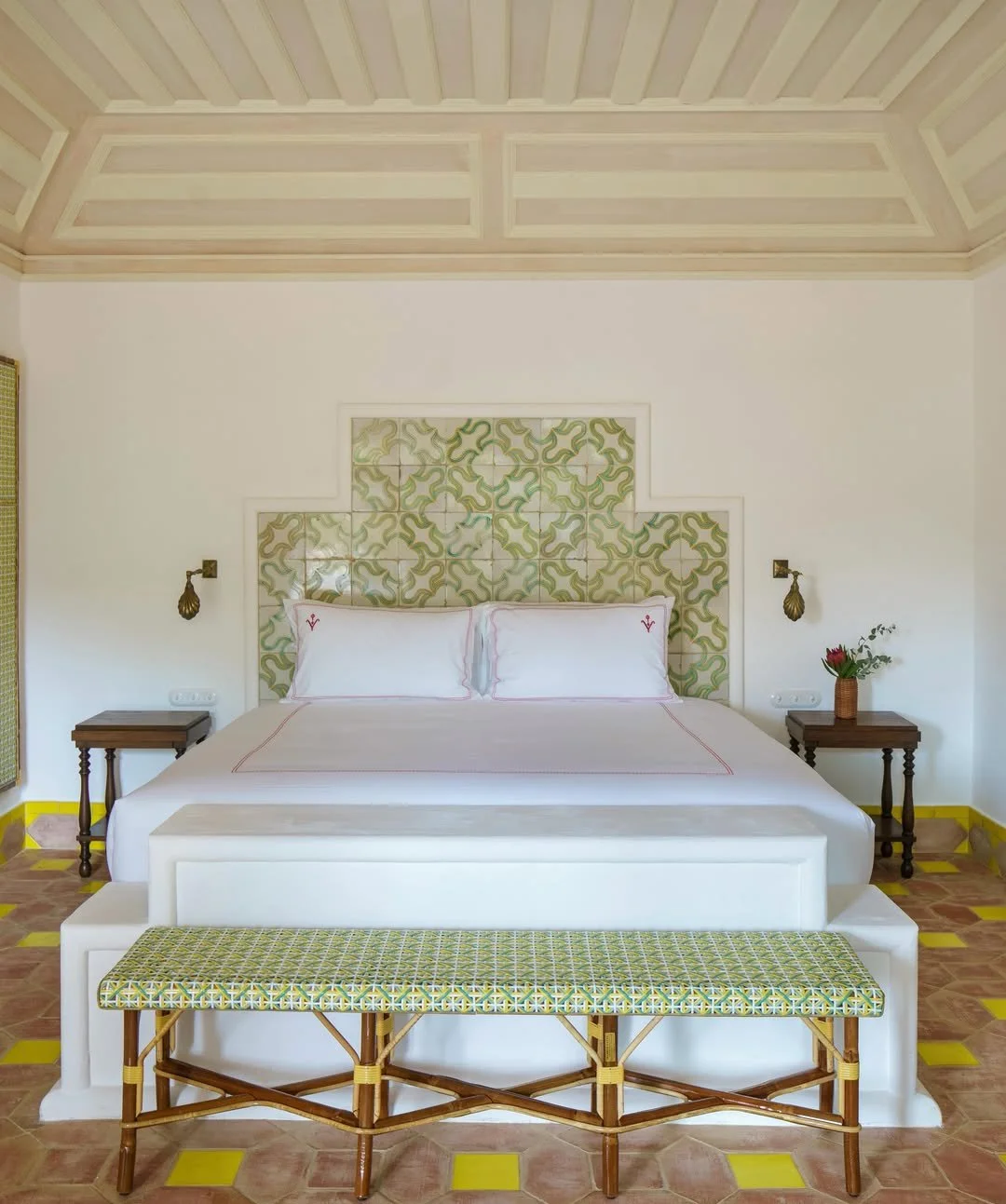 A bedroom with a white bed, a patterned headboard, two small wooden nightstands, wall-mounted lamps, a bench with a patterned cushion, a flower in a vase, and tiled flooring with yellow accents.