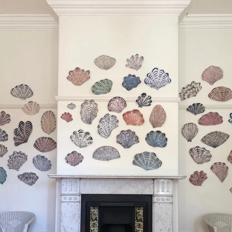 Interior of a room with white walls decorated with various woven shell-shaped wall hangings above a white marble fireplace mantel.