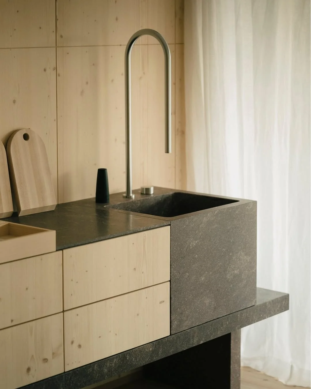 Minimalist kitchen sink with water faucet, black soap dispenser, and light wooden cabinetry against a wooden wall with a white curtain.