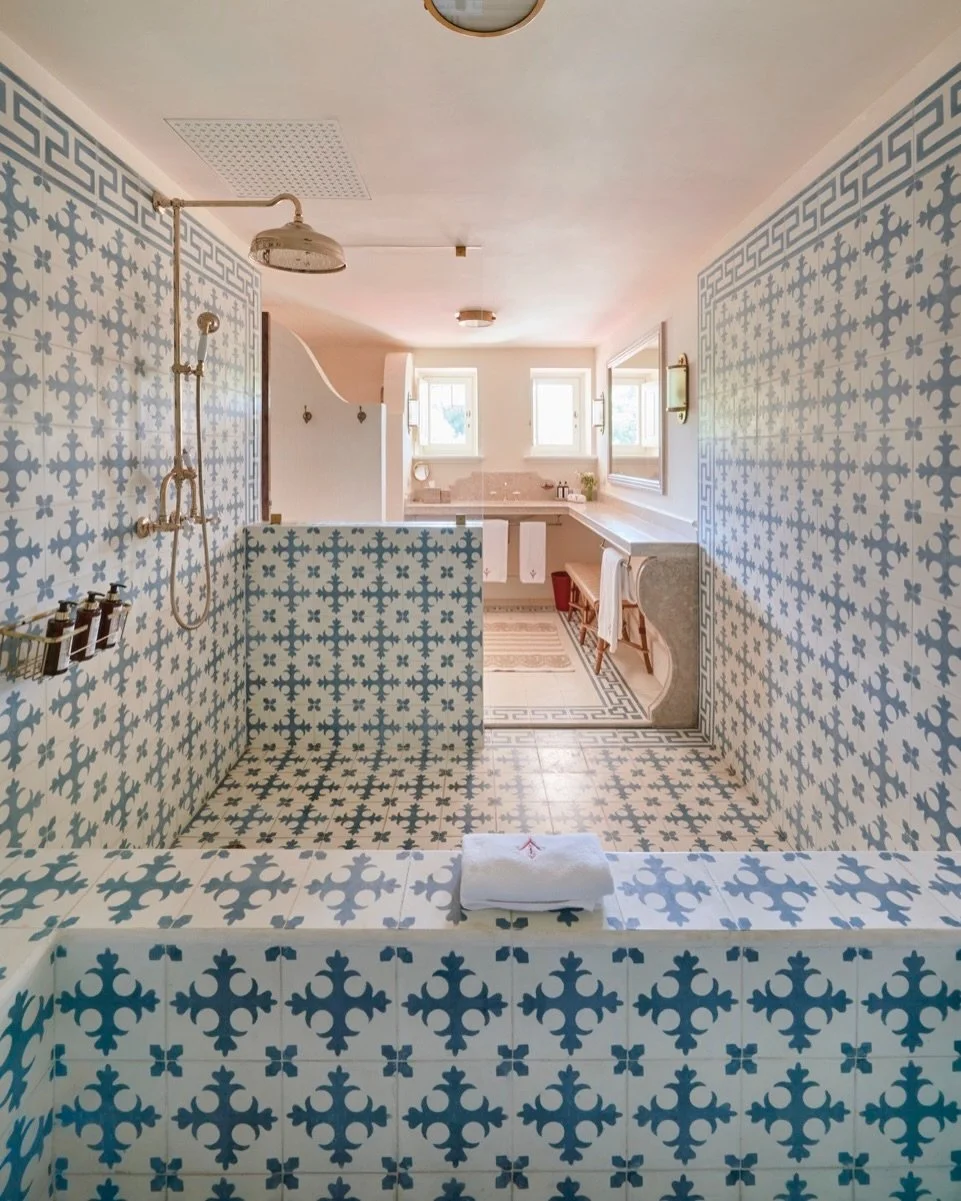 A spacious bathroom with blue and white patterned tiles on the walls, floor, and shower area, featuring vintage-style fixtures, a large window, a mirror, a countertop, and a bench.