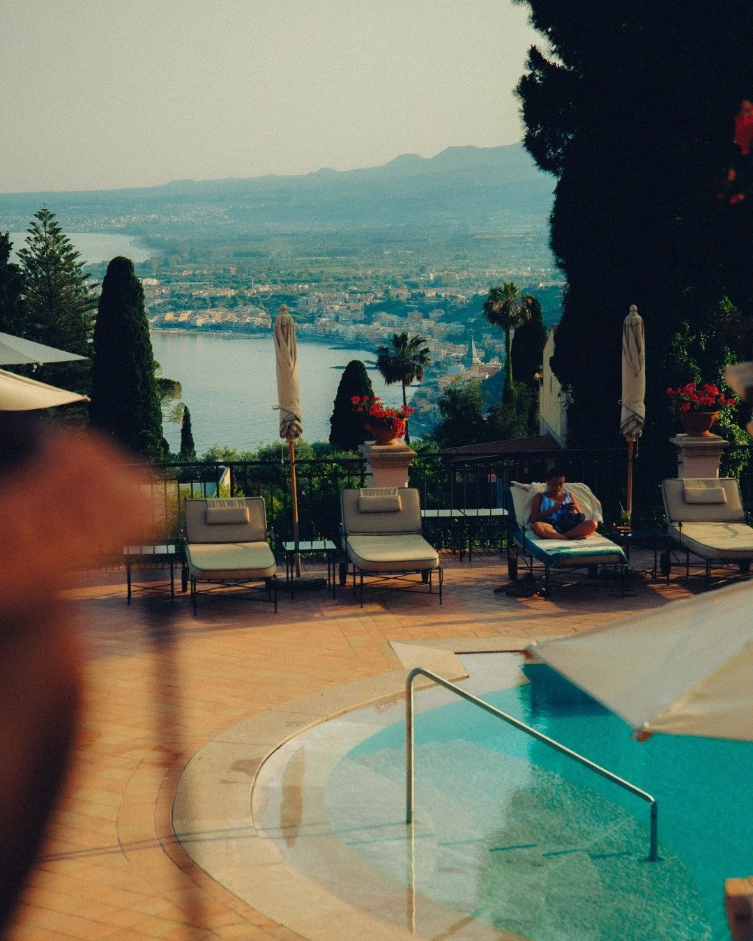 A swimming pool area with lounge chairs, umbrellas, and decorative plants overlooking a scenic city view with a large body of water and mountains in the distance.
