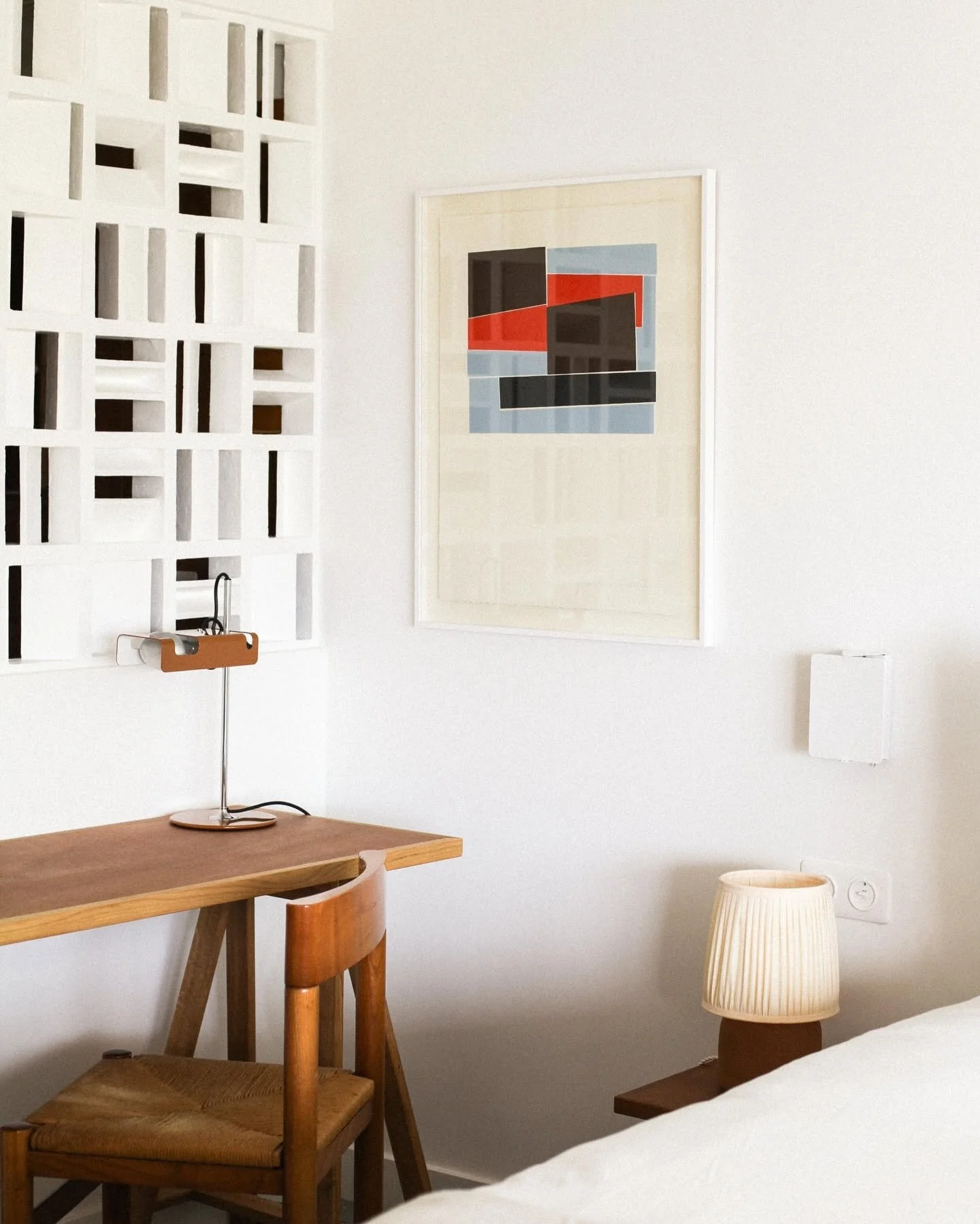 Interior view of a minimalist bedroom with a wooden table, a wooden chair with woven seat, a modern table lamp, a large abstract artwork on the wall, and a cream-colored bedside lamp.