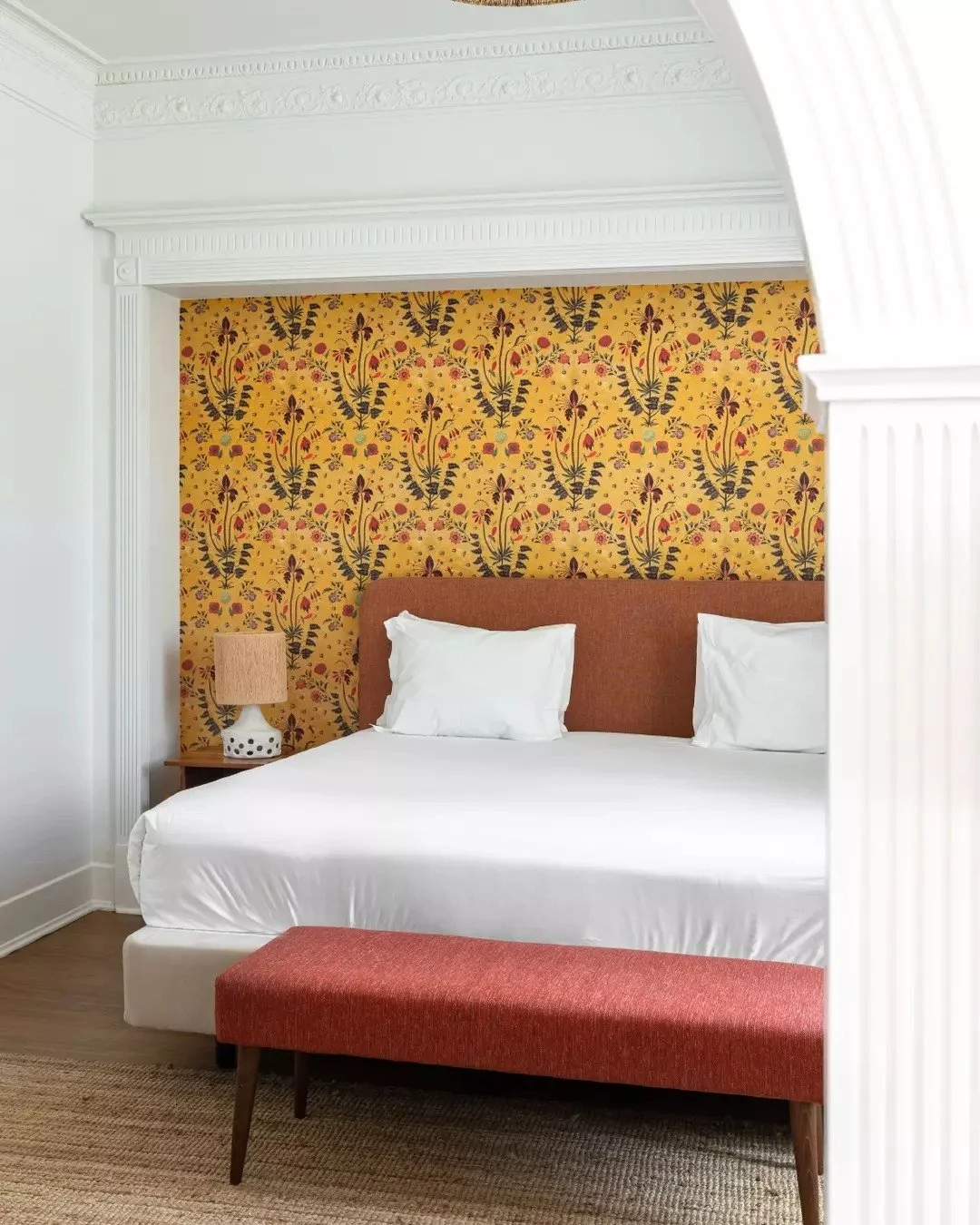A bedroom with a white bed, orange headboard, yellow floral wallpaper, bedside lamp, red bench, and white decorative moldings.