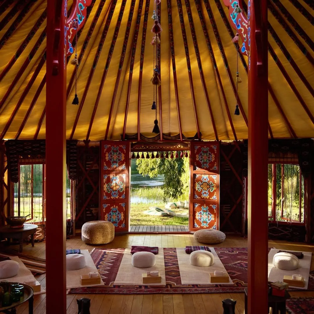 Interior of a tipi tent with a view of a lake and trees outside, with cushions and low tables on a rug inside.
