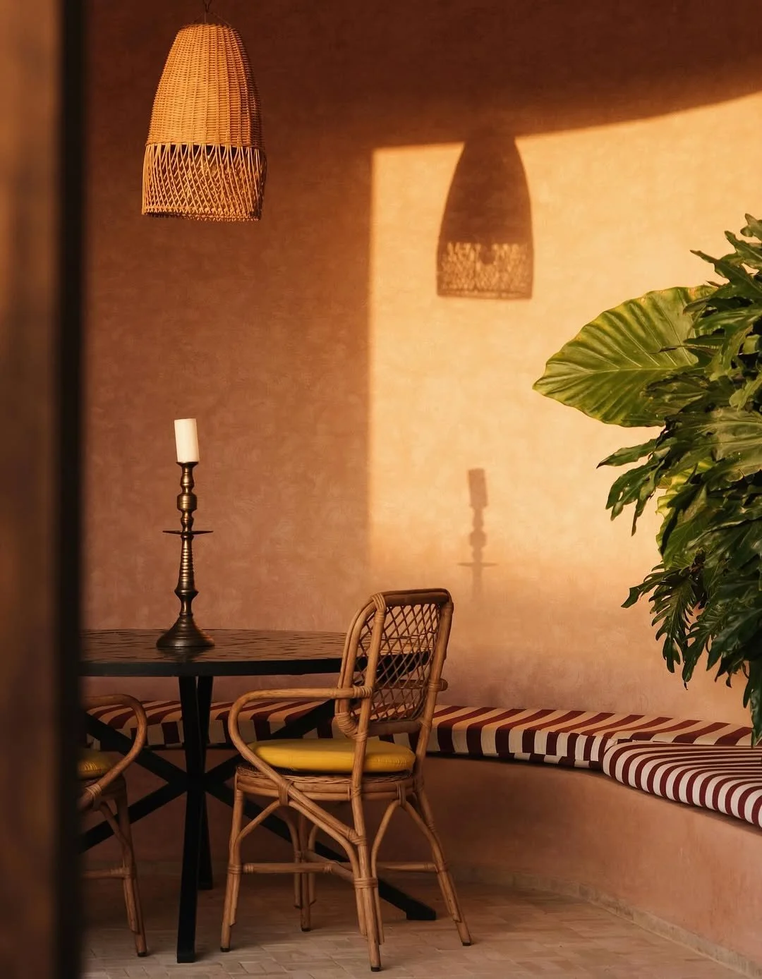 A cozy indoor space with warm lighting, a black table, rattan chairs with yellow cushions, a wall-mounted wicker pendant light, a candlestick with a white candle, striped cushioned seating, and a large green plant.