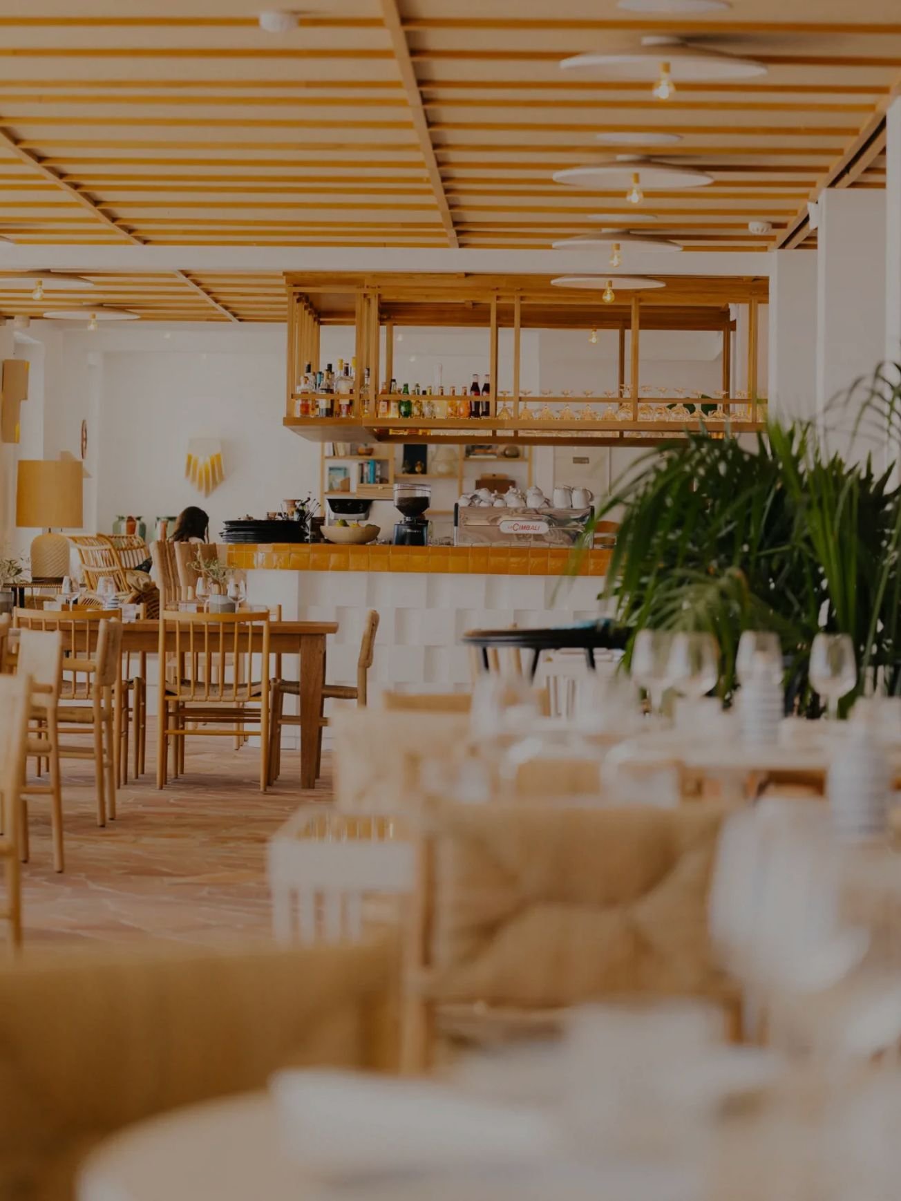 Interior of a restaurant with wooden tables and chairs, a bar area with bottles, and a person seated in the background.