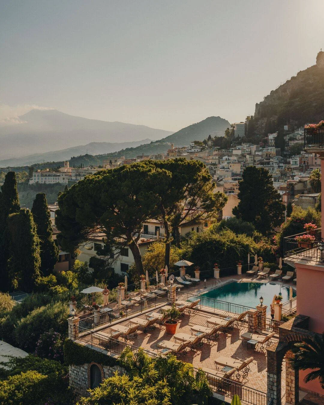 A scenic view of a hillside town with buildings, lush trees, and a swimming pool surrounded by sun loungers and umbrellas in the foreground, with mountains in the background.
