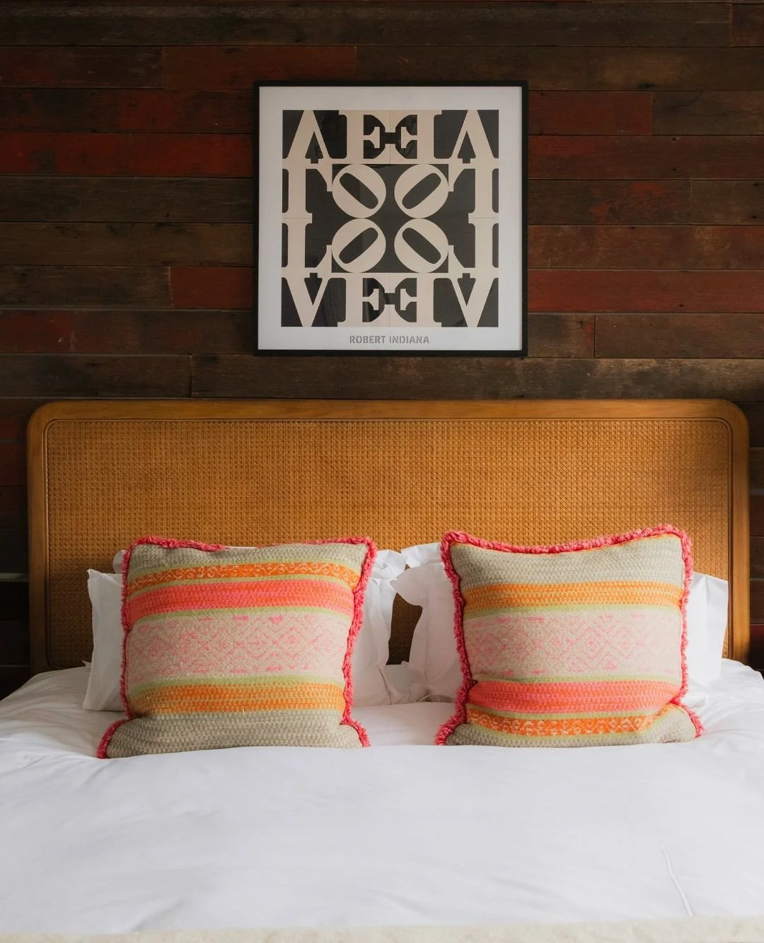 A neatly made bed with a wooden headboard featuring two decorative pillows with pink, orange, and green patterns and a fringed edge. Above the bed, a framed geometric art print with the word "LOVE" and a label with the name "Robert Indiana" is mounted on a dark wooden wall.
