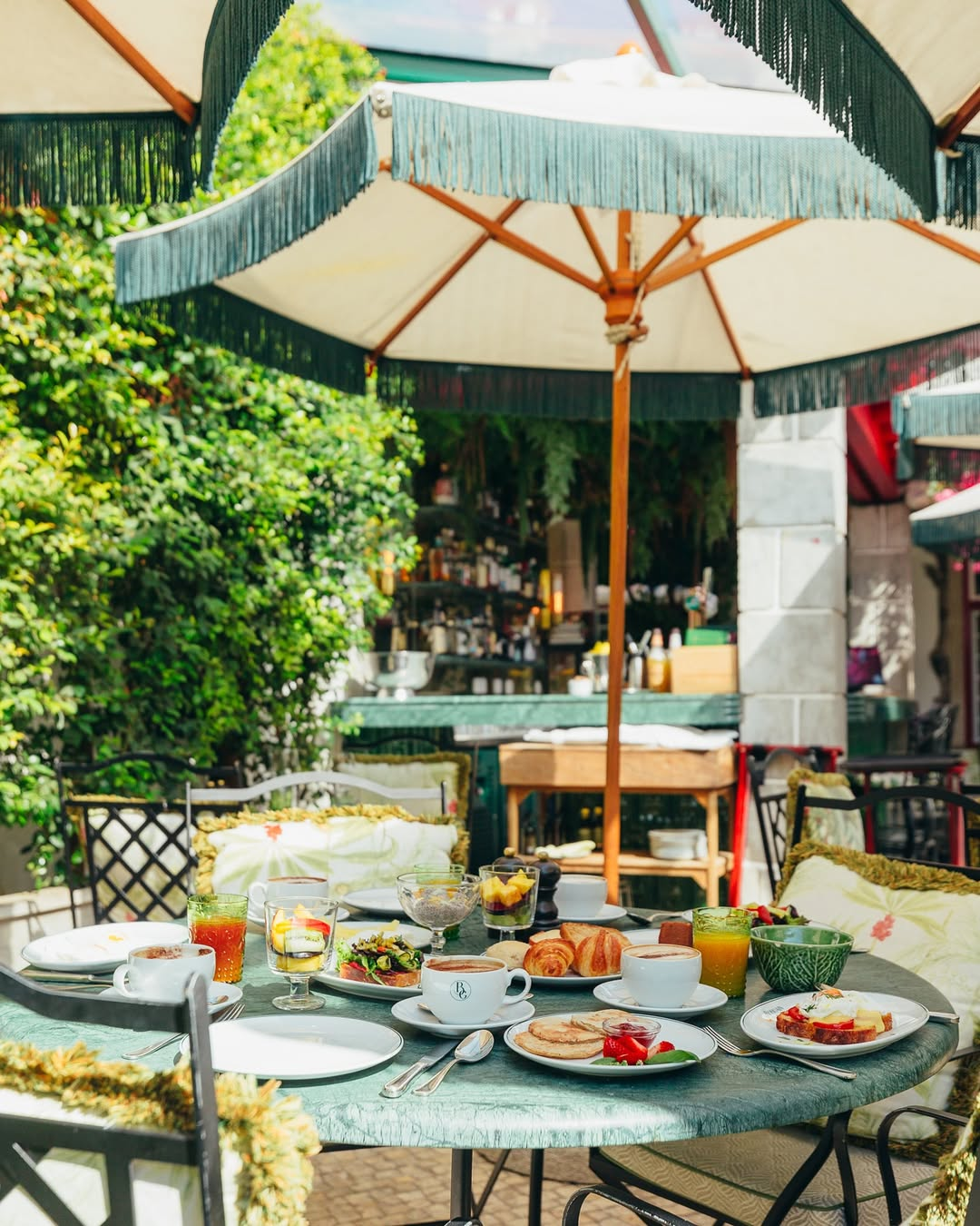 Outdoor breakfast table with various foods, drinks, and desserts under a large patio umbrella, surrounded by greenery.