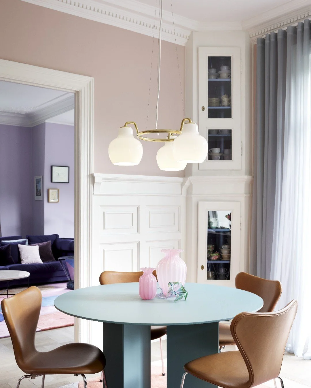 A dining room with a round table, pink vases, a white and gold hanging light, and a beige curtain. There is a living room with a purple sofa and picture frames on the wall visible in the background.